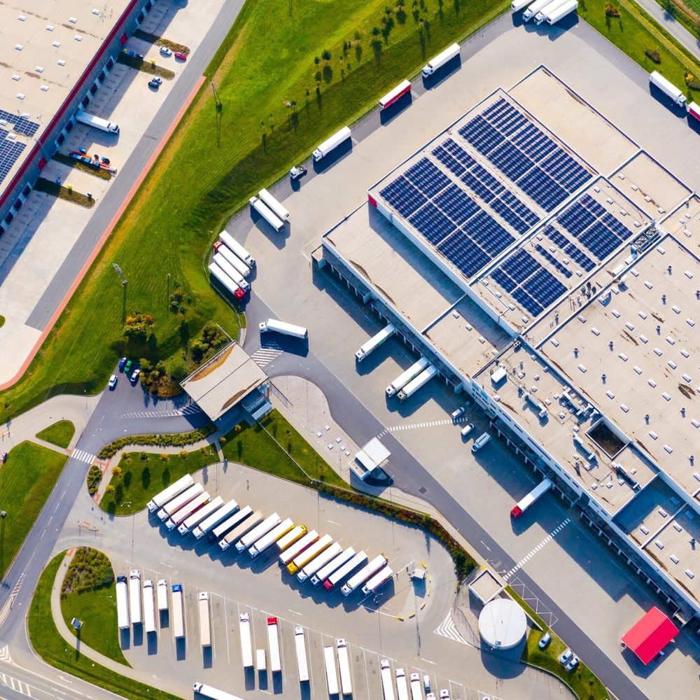 Industrihallar och logistikbyggnader – flygvy Flygbild över industrihallar och logistikbyggnader med lastbilar och solpaneler på taken.